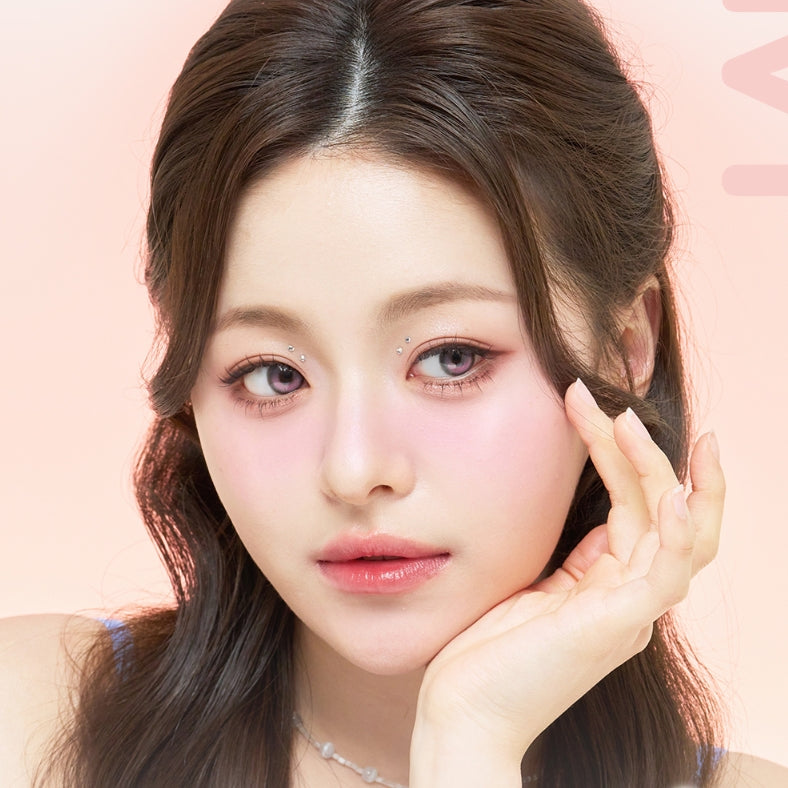A close-up portrait of a young woman with long dark brown hair, wearing soft pink makeup and a delicate silver necklace, posing with her hand against a pale pink background.
